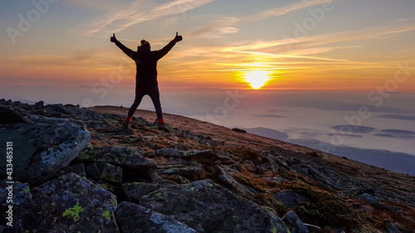Fototapeta A woman with a headband standing on top of Babia Gora, Poland, spreading her arms and enjoying the panoramic view on sun rising above the horizon. Thick clouds below. The sky is pink and orange. Free