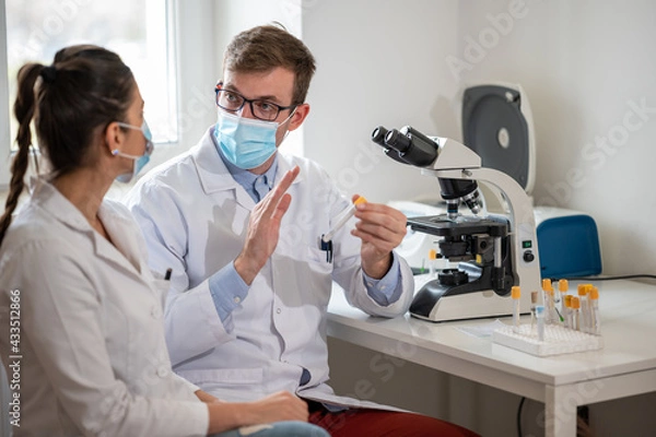 Fototapeta Two lab technicians with face masks working together in the laboratory and using a microscope.	