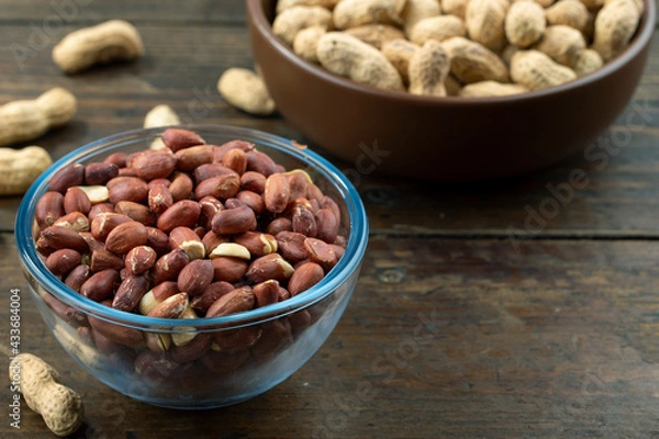 Fototapeta Peanuts in their husks in a glass bowl and peanuts in their skins are scattered on the table. Space for text.