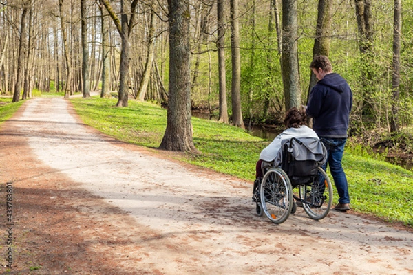 Obraz View from back to a woman in wheelchair and a man walking and checking his phone in a park near river at spring. Woman with a disability in wheelchair and her personal assistant are hiking together
