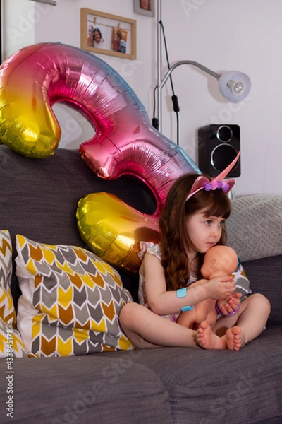 Obraz A Cute Baby Girl Sitting On A Sofa With A Colourful Balloon And A Doll