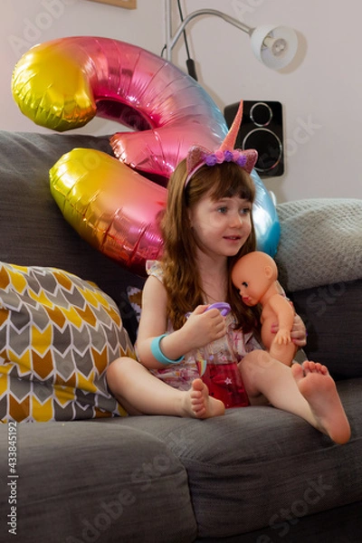 Obraz A Cute Baby Girl Sitting On A Sofa With A Colourful Balloon And A Doll