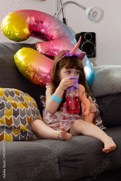 Obraz A Cute Baby Girl Sitting On A Sofa With A Colourful Balloon And A Doll