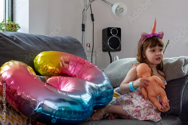 Obraz A Cute Baby Girl Sitting On A Sofa With A Colourful Balloon And A Doll