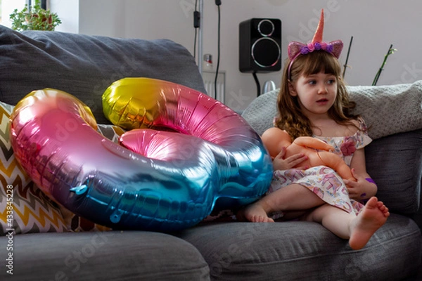 Obraz A Cute Baby Girl Sitting On A Sofa With A Colourful Balloon And A Doll