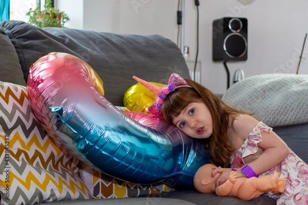 Obraz A Cute Baby Girl Sitting On A Sofa With A Colourful Balloon And A Doll