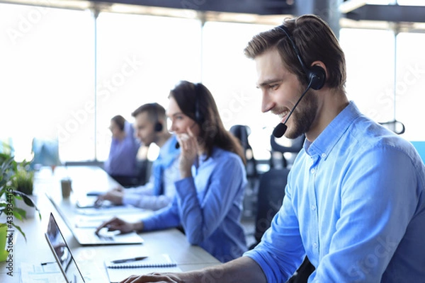 Fototapeta Smiling male call-center operator with headphones sitting at modern office with collegues on the backgroung, consulting online.