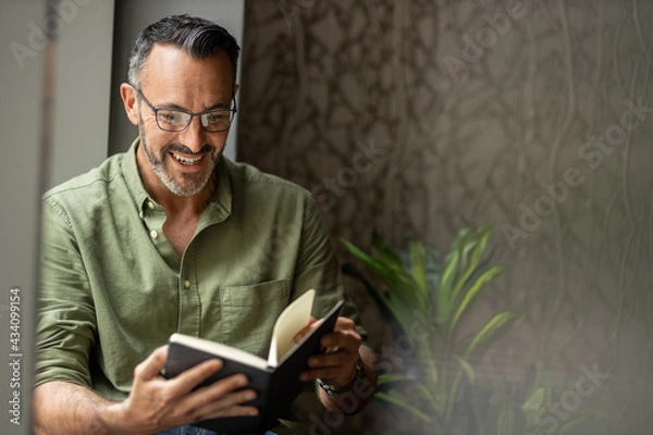 Fototapeta Mature man reading book by window, smiling wearing glasses, copy space