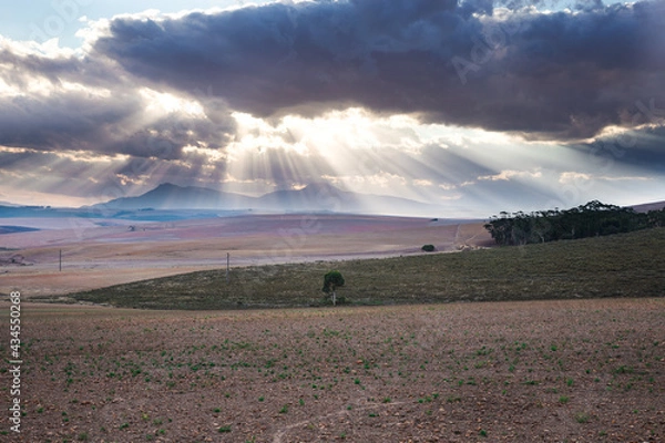 Fototapeta Sunrays burst through clouds over a mountain with farm land in the foreground