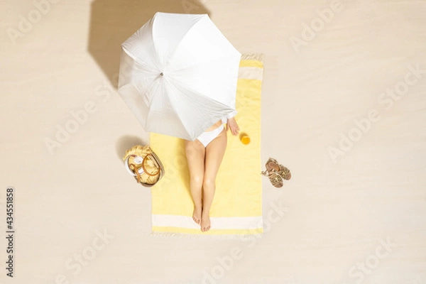 Fototapeta Alone woman in bikini sitting and sunbathes on yellow beach towel under umbrella with bag and juice. Female relaxation on the sand beach at summer vacation. Top, aerial view