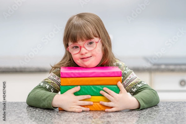 Fototapeta A girl with down syndrome sitting at a table at home, leaning on a stack of books that are in front of her, looking at the camera with smile