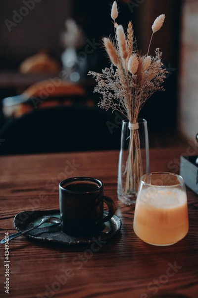 Obraz A cup of black coffee, orange juice and dried flowers in a glass vase standing on a wooden table in the interior of a cafe