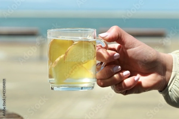 Obraz Ginger tea with slices of ginger hold by a hand in front of a beach