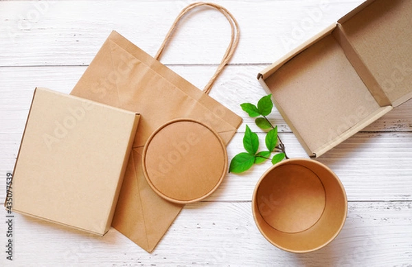Fototapeta Top view of Paper box packaging in rectangle and round shape and paper bag with green leaves on white wooden background , delivery eco package and save environment  concept