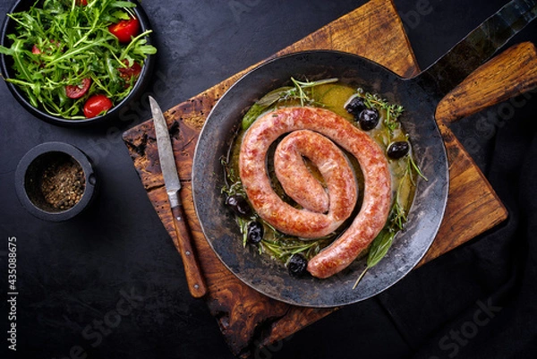 Fototapeta Traditional fried Italian salsiccia fresco meat sausage served with rocket salad and olives as top view in a cast iron pan