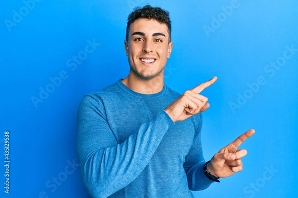 Fototapeta Hispanic young man wearing casual winter sweater smiling and looking at the camera pointing with two hands and fingers to the side.