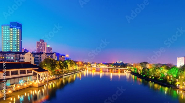 Fototapeta Long exposure shot at blue hours with flare of lamp light from riverfront restaurant, bridge light and building in the evening time, Landscape and city scape in high saturation tone.