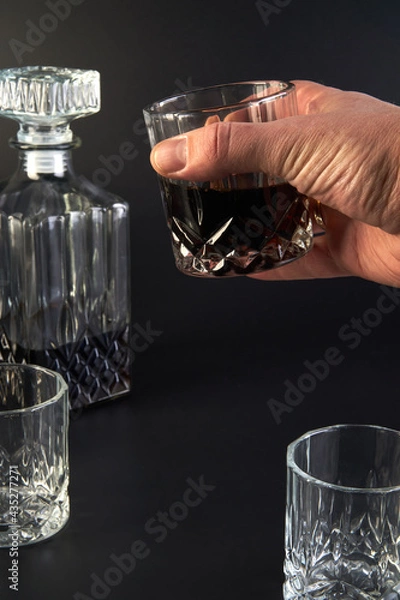Fototapeta A man holds a glass of cognac in his hand next to other empty glasses and a decanter for cognac and whiskey on a dark background. Cognac is in the decanter. Close-up