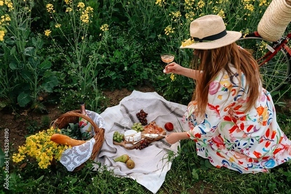 Fototapeta A beautiful girl drinksing wine at a picnic in a field of flowers. A summer eco picnic in the field with straw basket, flowers, wine, fruits, baguette, cheese Camembert