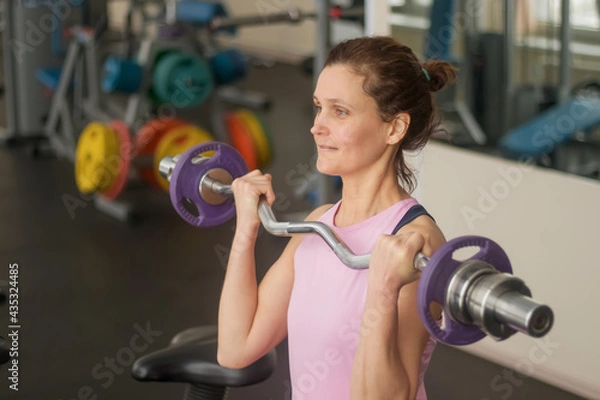 Obraz A 40-45 woman does a bicep exercise with a barbell in the gym