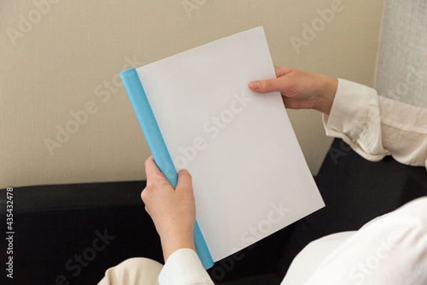 Fototapeta Beautiful female hands hold an open book or magazine in a room on a black sofa
