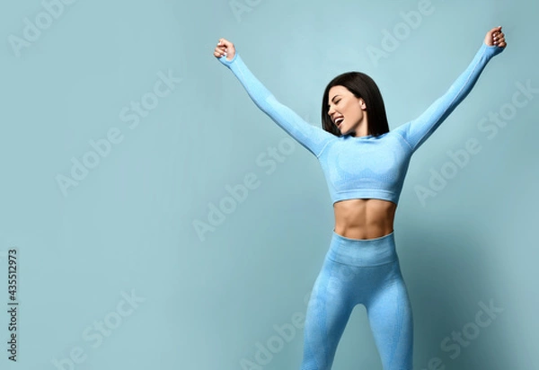 Fototapeta Portrait of fitness girl. Woman in fashion sportswear, standing on a blue background, after a workout. Fit girls with a strong muscular body, raised her arms up, rejoices in her body