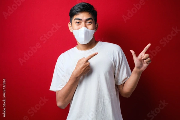 Fototapeta Young handsome man wearing medical mask smiling and looking at the camera pointing with one hands and fingers to the side