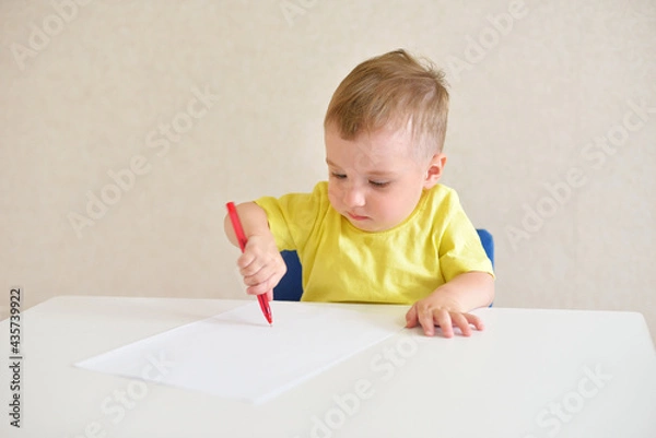 Fototapeta toddler boy plays draws on a piece of paper with a red pen sitting at a white table. educational game Montessori fine motor skills. early development concept.