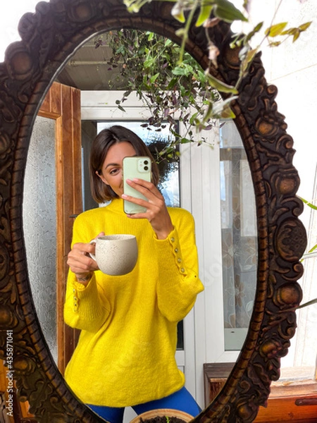 Fototapeta Pretty woman at home take photo selfie in mirror on mobile phone for stories and posts in social media, vertical frame, wearing cosy comfy warm yellow sweater hold mug