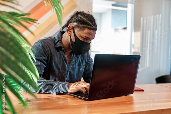 Obraz young latin man sitting at home working from his living room using laptop with mask and quarantine