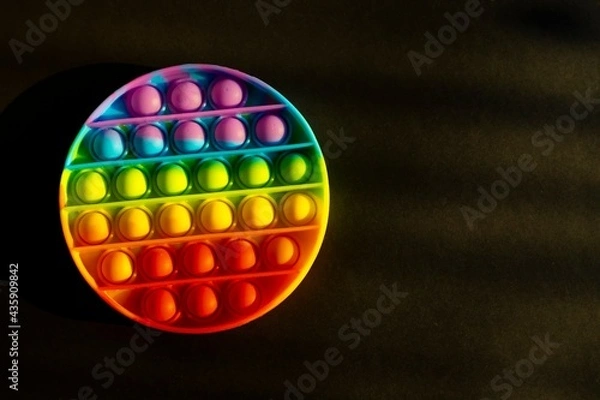 Fototapeta Trendy colorful anti-stress sensory toy. Silicone fidget push pop it for fine motoric skills lies in the sun. Flat lay on black background. Picture with place for text.