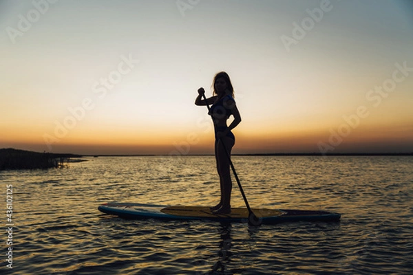 Obraz Young beautiful girl in a bikini is resting on a surfboard at sunset