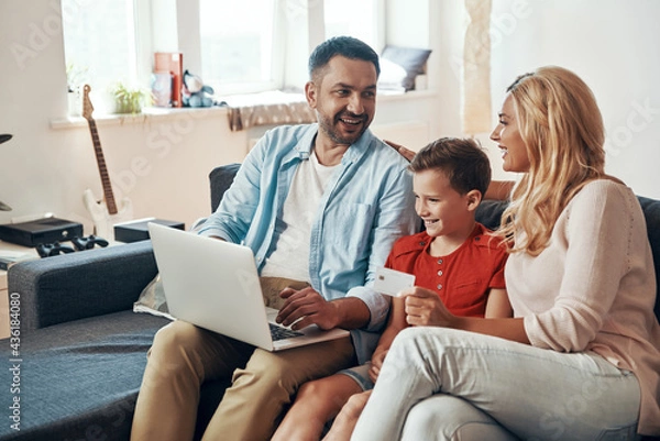 Obraz Young beautiful family shopping online and smiling while using laptop at home