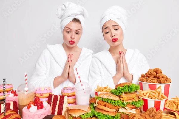Fototapeta Sorrowful diverse women with pleading expressions keep palms pressed together look at appetizing junk food feel temptation to eat something delicious and harmful for your health dressed in bathrobe