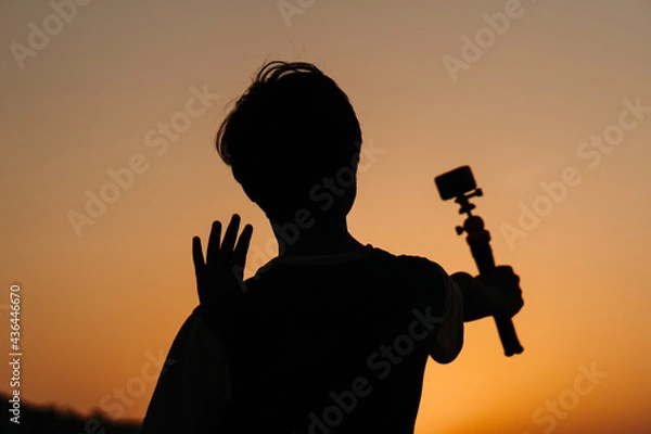 Fototapeta Silhouette of an Indian teen vlogging while holding camera in his hand. Teenager vlogging for social Media. Young teen holding camera and speaking at the camera