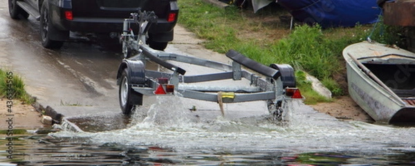Fototapeta Motor boat launch, SUV car with wet empty boat trailer move on concrete shipway from water at summer day on old boat on shore background