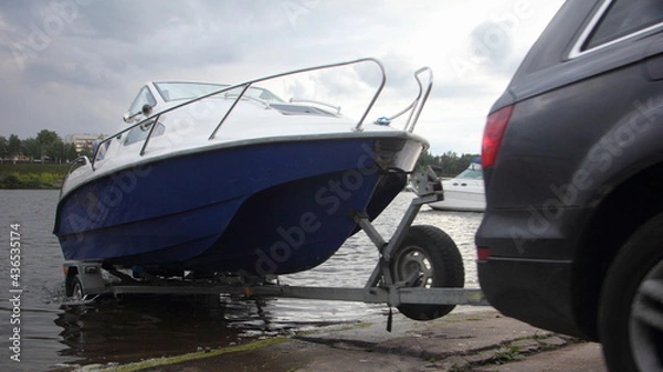 Fototapeta A 4x4 black car pulls blue-white trimaran cabin motor boat on trailer on concrete slipway into the river water, boat launch before active recreation on the river