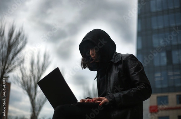 Fototapeta Girl in black leather coat and hood with laptop on city street, concept of woman hacker and agent working remotely at computer