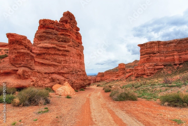 Obraz Canyon, mountain erosion, plateau, cloudy sky and mountains on the horizon. Red rocks and layers. The road along the bottom of the canyon. Charyn canyon in Kazakhstan