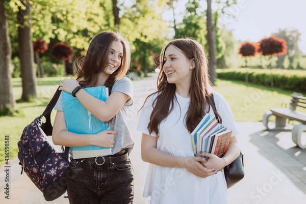 Fototapeta Two happy smiling student girls are walking and talking to each other on campus on a warm sunny day. Soft selective focus.