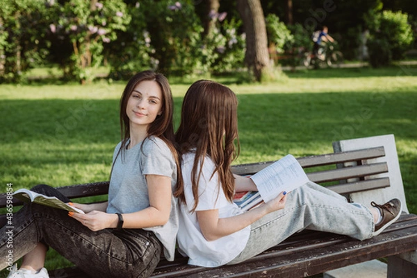 Fototapeta Two student girls are looking at an open book on a bench in the park. Distance education, preparation for exams. Soft selective focus.