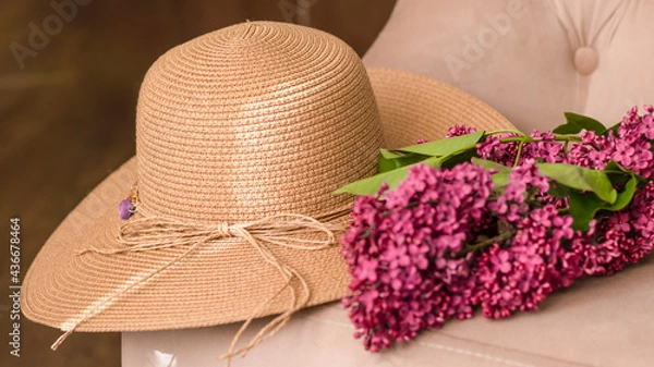 Fototapeta Decorative composition with a bouquet of lilacs and a beige wicker hat. Beach wicker hat, flowers, close-up. Lilac bouquet. Cozy atmosphere of relaxation and romance. Summer background. 