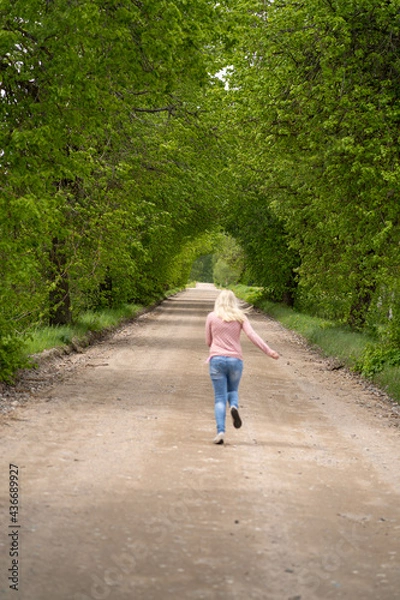 Fototapeta The girl blonde on the road runs in a tree arch