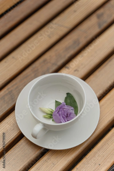 Obraz A white cup and saucer stand on a wooden background. There is a purple flower in the cup. Flower tea. Drink tea outside. Romantic mood.