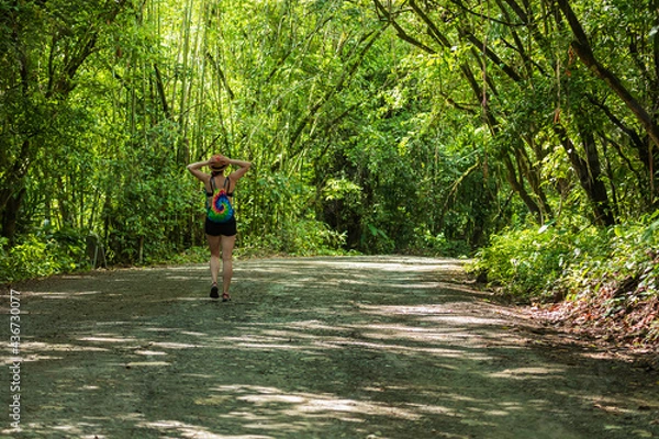 Fototapeta Woman with colorful backpack walking on rural road through tropical forest with copy space