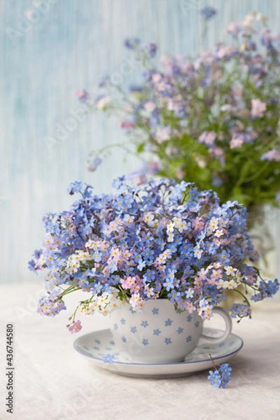 Obraz A bouquet of spring flowers forget-me-nots in a cup on the table against the background of a decorative colored wall. Postcard, blur, selective focus.