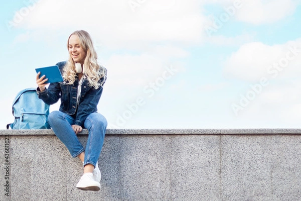 Fototapeta Smiling young female student is studying on blue sky background. Blond girl is preparing to exam, holding tablet computer at college university campus. High school lifestyle. Education concept.