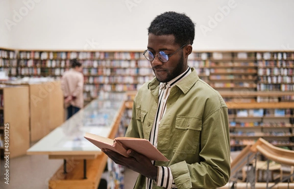 Obraz African young man in eyeglasses reading a book while standing in the library