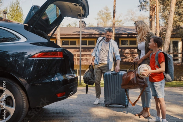 Fototapeta Happy young family with little packing stuff into the car and smiling while standing near house