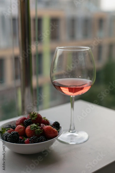 Obraz A glass of rose wine is on the table on the balcony, next to a plate of berries. Drink wine and eat berries. The mood of summer. Wine with fruit. Summer vacation with wine. Alcoholic beverages.